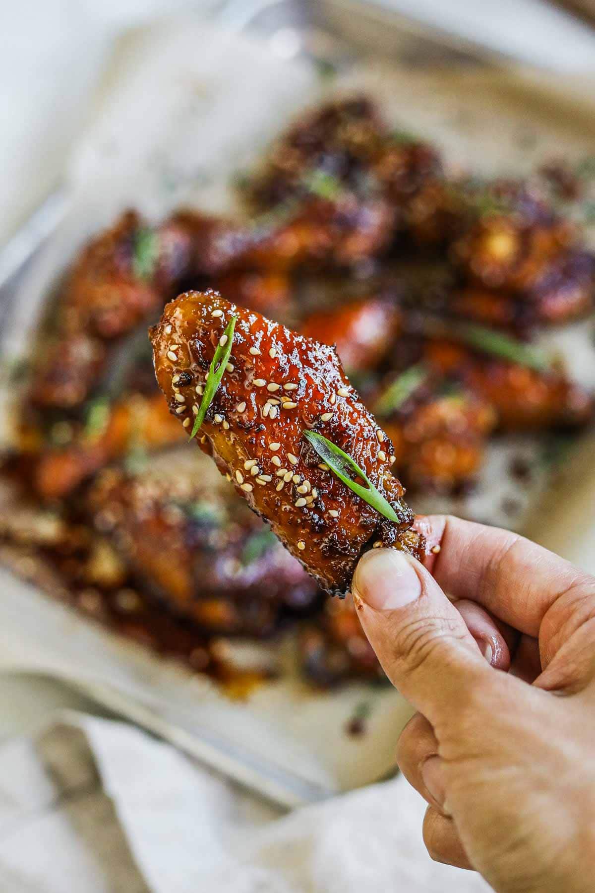 Hand holding a honey soy garlic flat wing with sliced green onion and sesame seeds.
