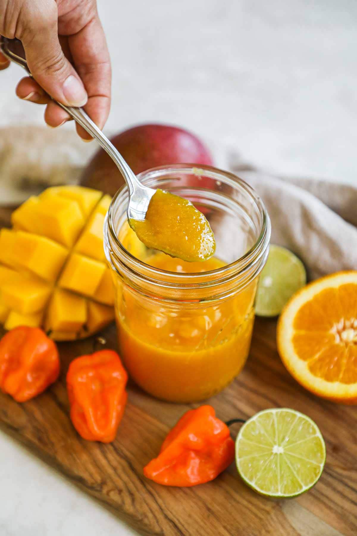 Hand holding spoon of mango habanero sauce in a jar with mango, habanero peppers, lime, and orange.