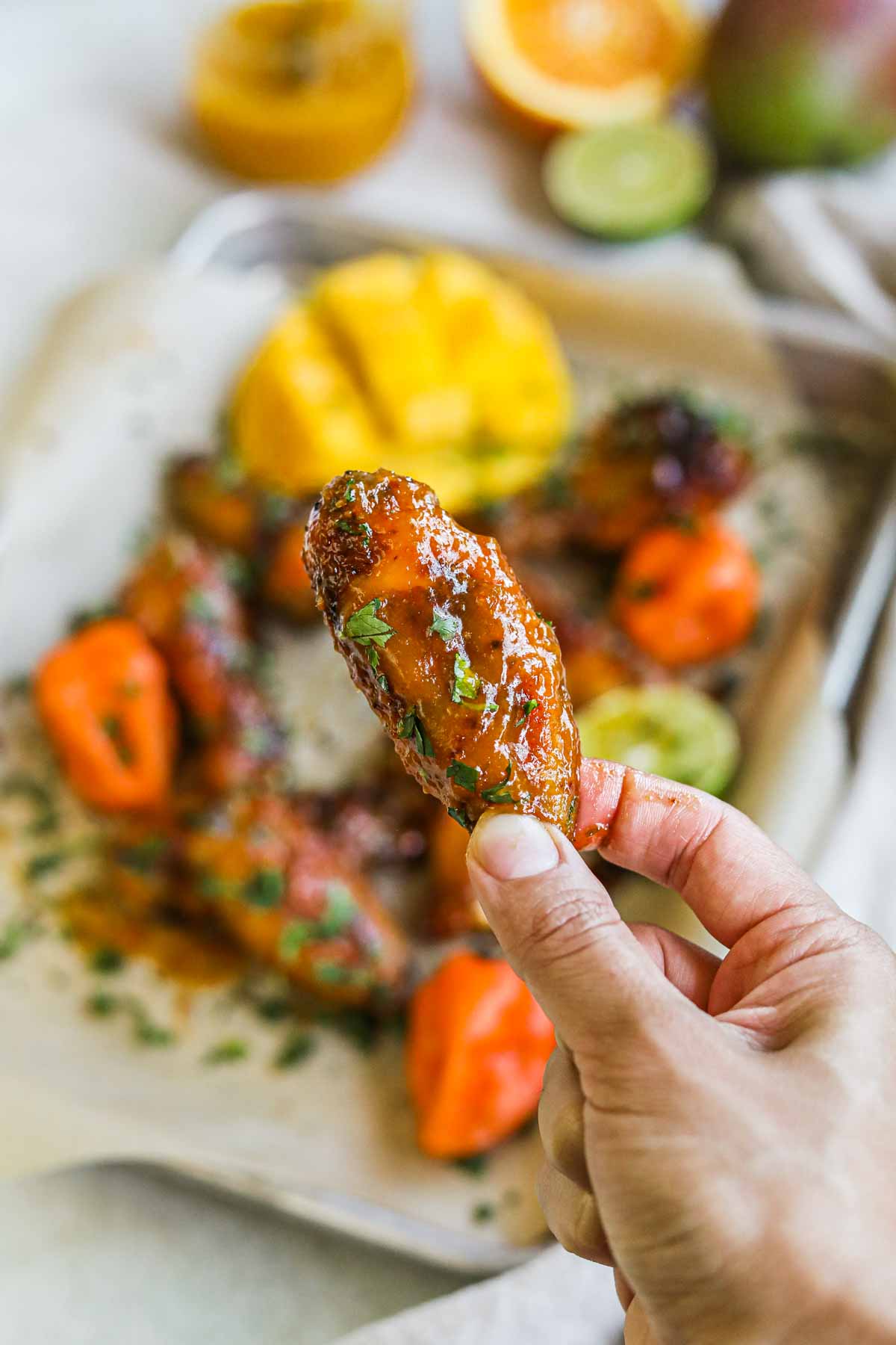 Hand holding a sticky, sweet, and spicy mango habanero chicken wing above a platter of wings.