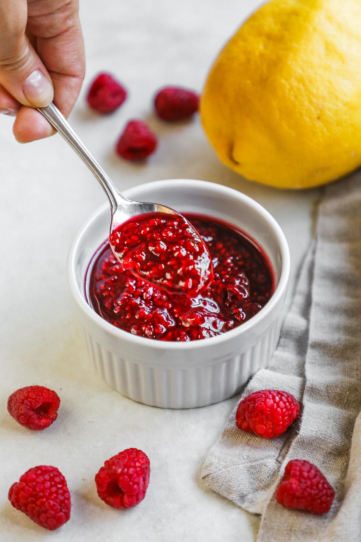 Spoon holding a scoop of handmade raspberry compote above a ramekin of compote for ice cream, cake, cheesecake, dessert, and more.