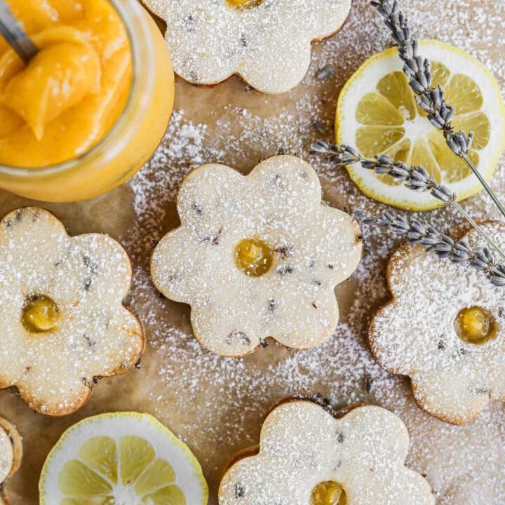 Flower lavender lemon curd cookies dusted with powdered sugar on parchment paper with lemon slices, lavender, and a jar of homemade lemon curd.