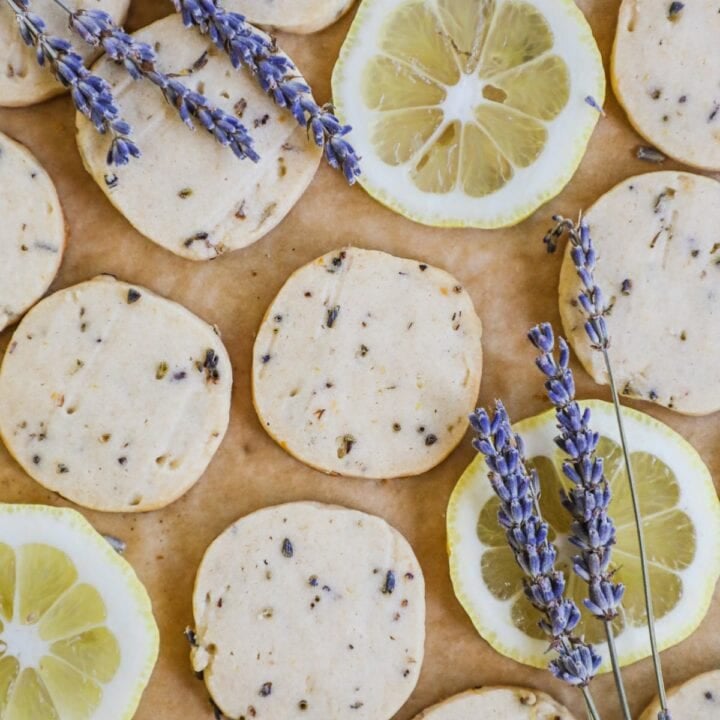 Rolled and sliced lemon lavender shortbread cookies on a sheet of parchment paper with lemon slices and sprigs of lavender.
