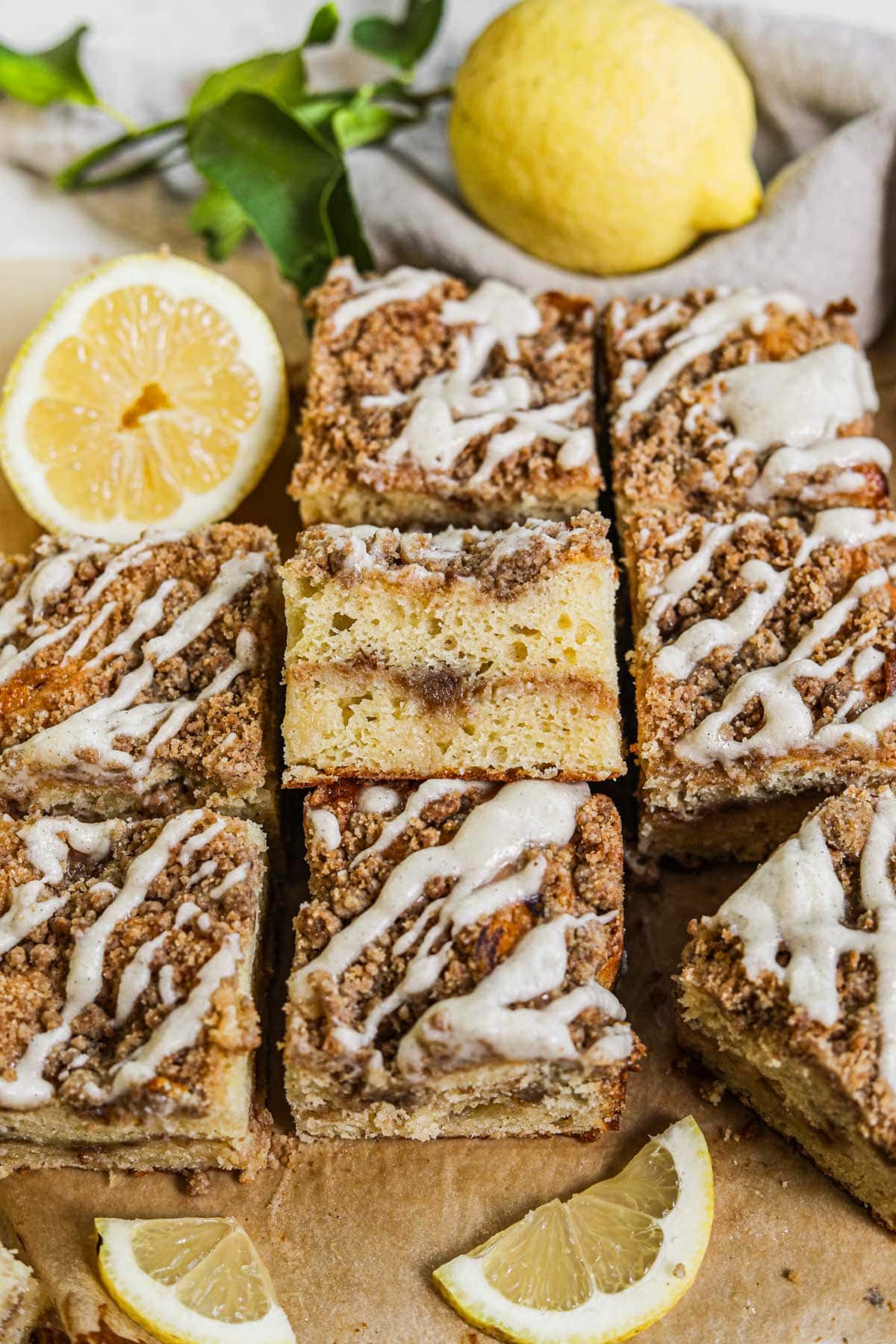 Sliced lemon curd coffee cake squares with cinnamon streusel and vanilla bean glaze drizzled on top, surrounded by sliced lemons.