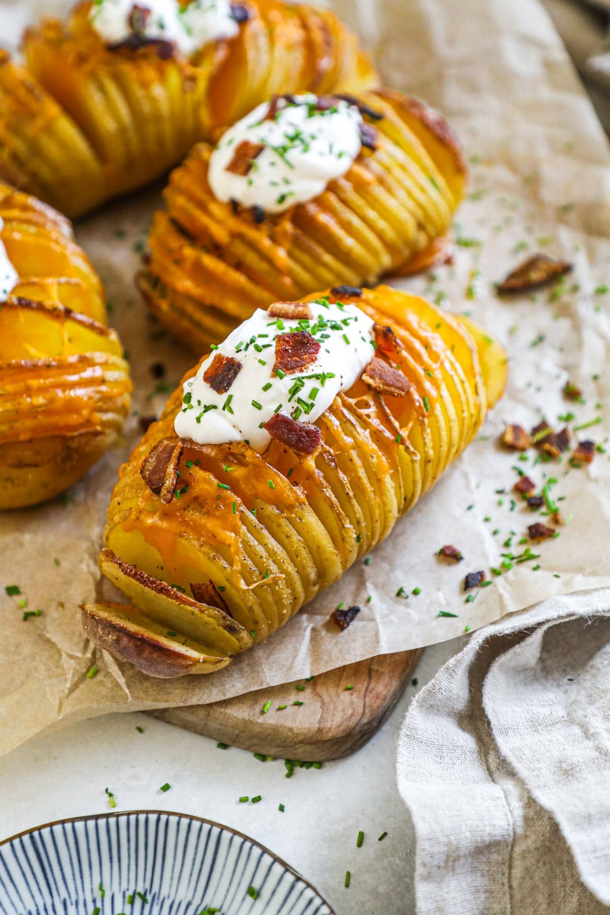 Bacon cheddar hasselback potatoes topped with sour cream, bacon, and chives on a parchment paper-lined board for serving.