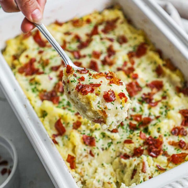 Creamy twice-cooked mashed potatoes with roasted garlic, gruyere, chives, and crispy prosciutto in a Le Creuset baker with a spoon holding a cheesy scoop.