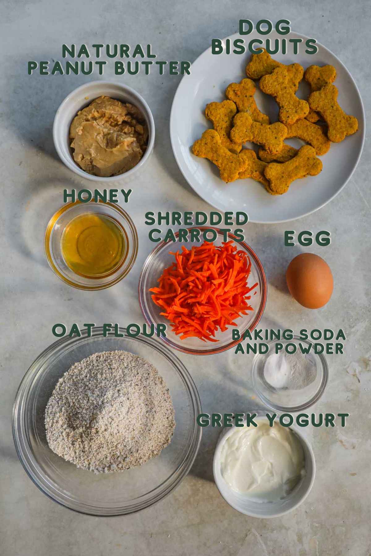 Ingredients to make pupcakes, shredded carrots, natural peanut butter, honey, oat flour, egg, baking soda, baking powder, greek yogurt, dog biscuits.