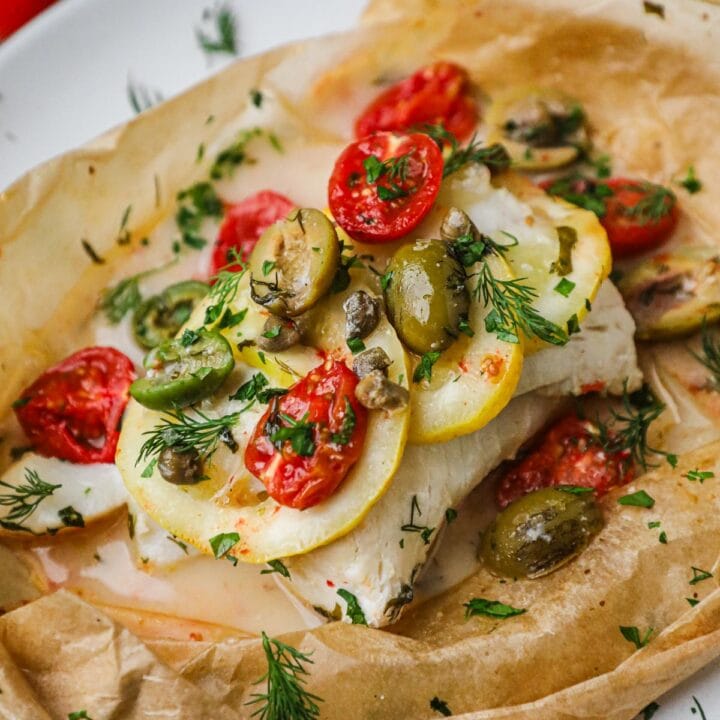 Baked cod in parchment paper (cod en papillote) with lemon, olives, tomatoes, capers, and fresh parsley.