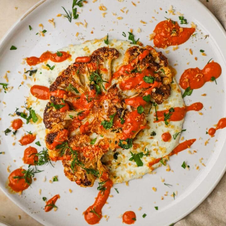 Roasted cauliflower steaks on a bed of whipped lemon feta, topped with a drizzle of harissa tahini sauce, herbed pangrattato, and fresh herbs,