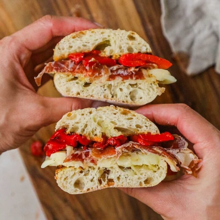 Hands holding capicola, provolone, roasted red peppers, and toasted ciabatta panino cut in half.