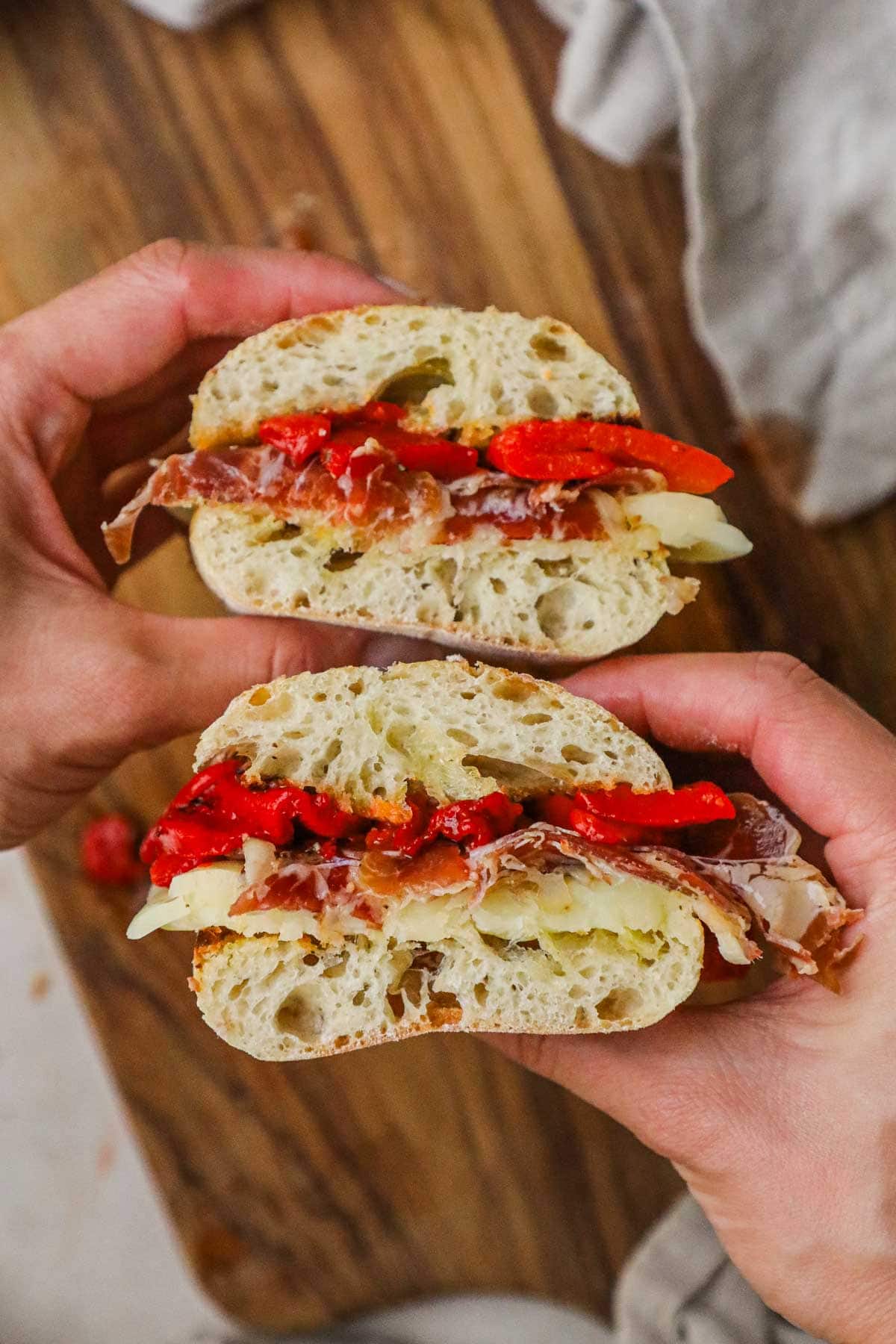 Hands holding capicola, provolone, roasted red peppers, and toasted ciabatta panino cut in half.