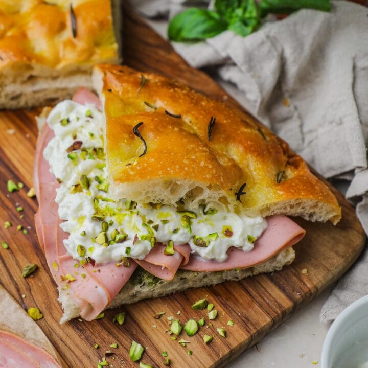 Authentic Italian panino made with focaccia, pesto, sliced mortadella, creamy burrata, crushed pistachios, olive oil, and flaky sea salt on an olive wood serving board.