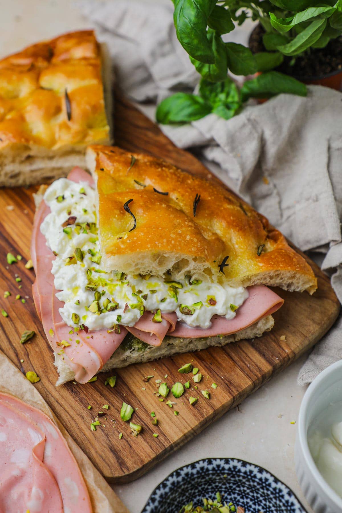 Authentic Italian panino made with focaccia, pesto, sliced mortadella, creamy burrata, crushed pistachios, olive oil, and flaky sea salt on an olive wood serving board.