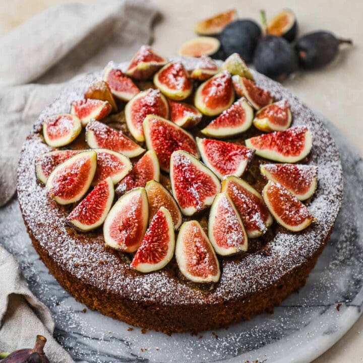 Fig and almond olive cake topped with fresh sliced figs, confectioners' sugar, and honey on a marble cake stand.